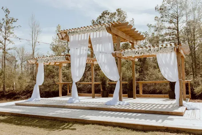 Outdoor wooden wedding arch with white drapes and fabric panels on a raised platform in a wooded setting