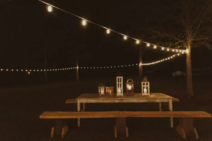 String lights over an outdoor picnic table at night, with lanterns on the table and bare trees in the background