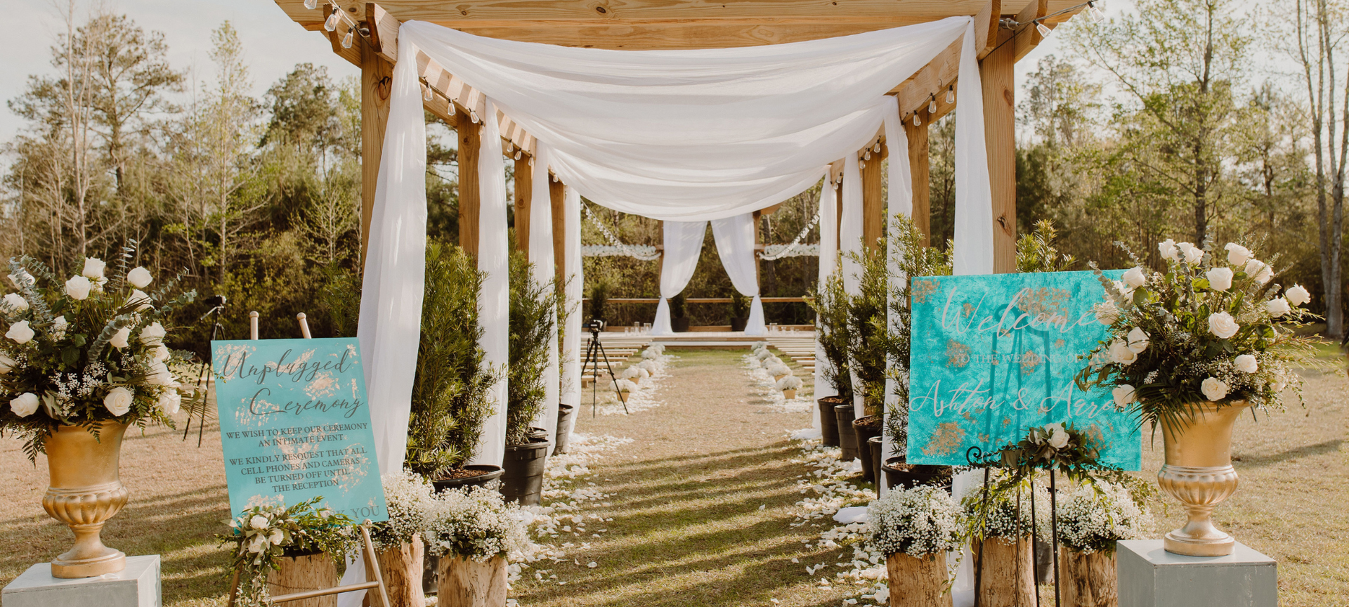 Outdoor wedding altar with white drapery, gold chairs, floral arrangements, and turquoise signs
