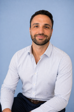 Smiling man in a white patterned shirt seated against a light blue background.