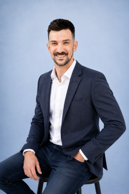 Man seated on a stool against a light gray background, wearing a beige sweater and navy pants, smiling at the camera