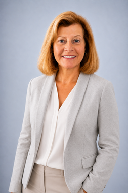 Man seated on a stool against a light gray background, wearing a beige sweater and navy pants, smiling at the camera
