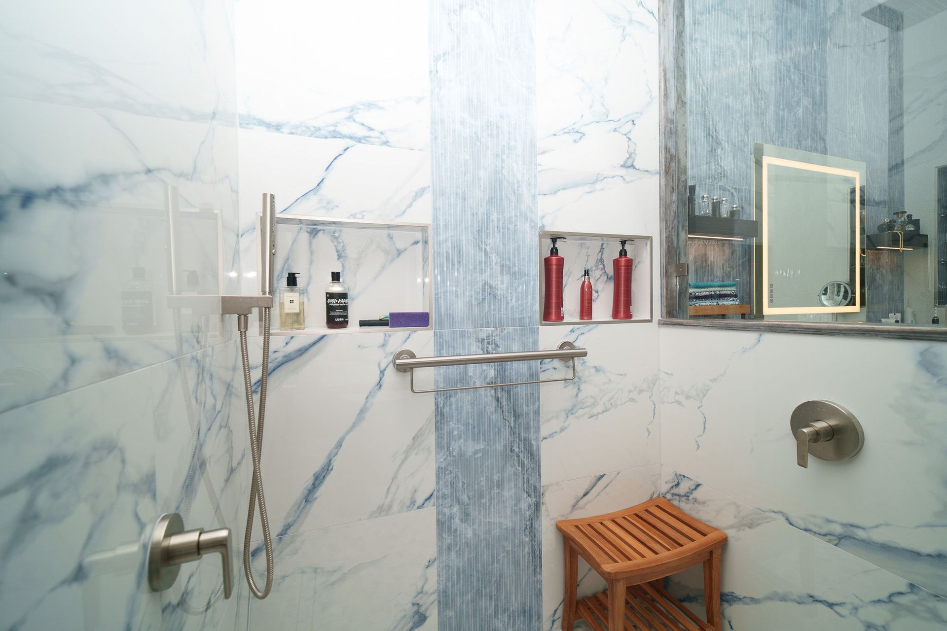 Modern bathroom with marble-patterned walls, glass shower, wooden stool, and toiletries on a shelf