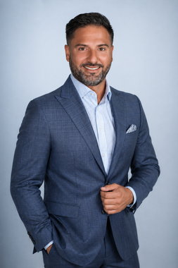 Portrait of a smiling man in a blue suit with a patterned shirt and pocket square against a light gray background