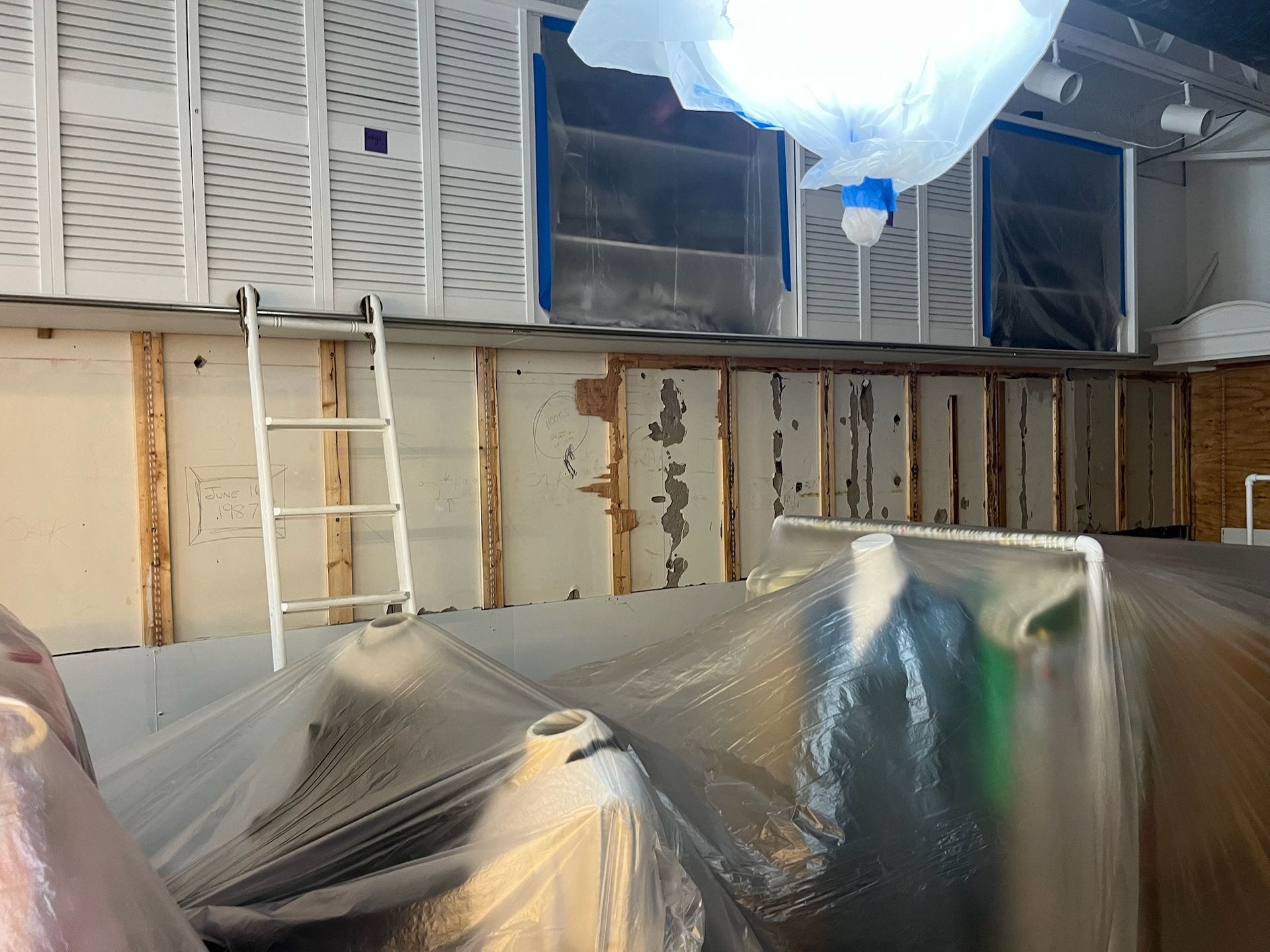 Construction site with plastic-covered equipment, a ladder, and exposed wall panels inside a room under renovation