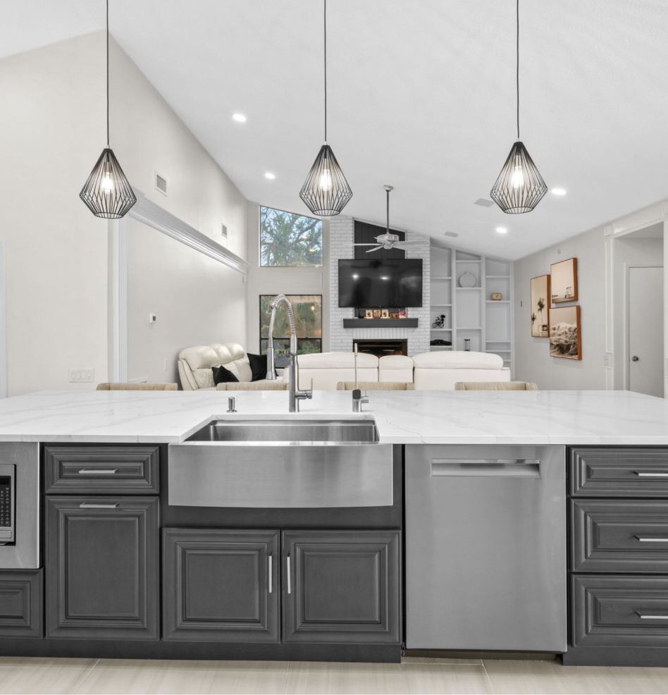 Modern kitchen with dark cabinets, white island, stainless sink, pendant lights, and open living room beyond