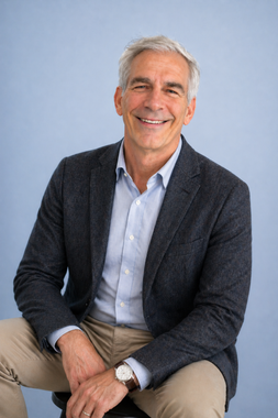 Man in a gray shirt with arms crossed, standing against a light blue background.