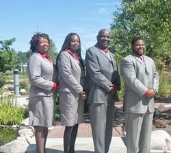 Four people in gray suits standing outdoors in a garden, smiling at the camera.