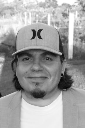 Man in a baseball cap and blazer smiling outdoors in black and white.