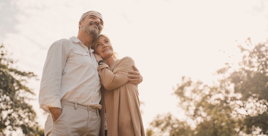 A couple embracing outdoors under a bright sky, surrounded by soft trees.