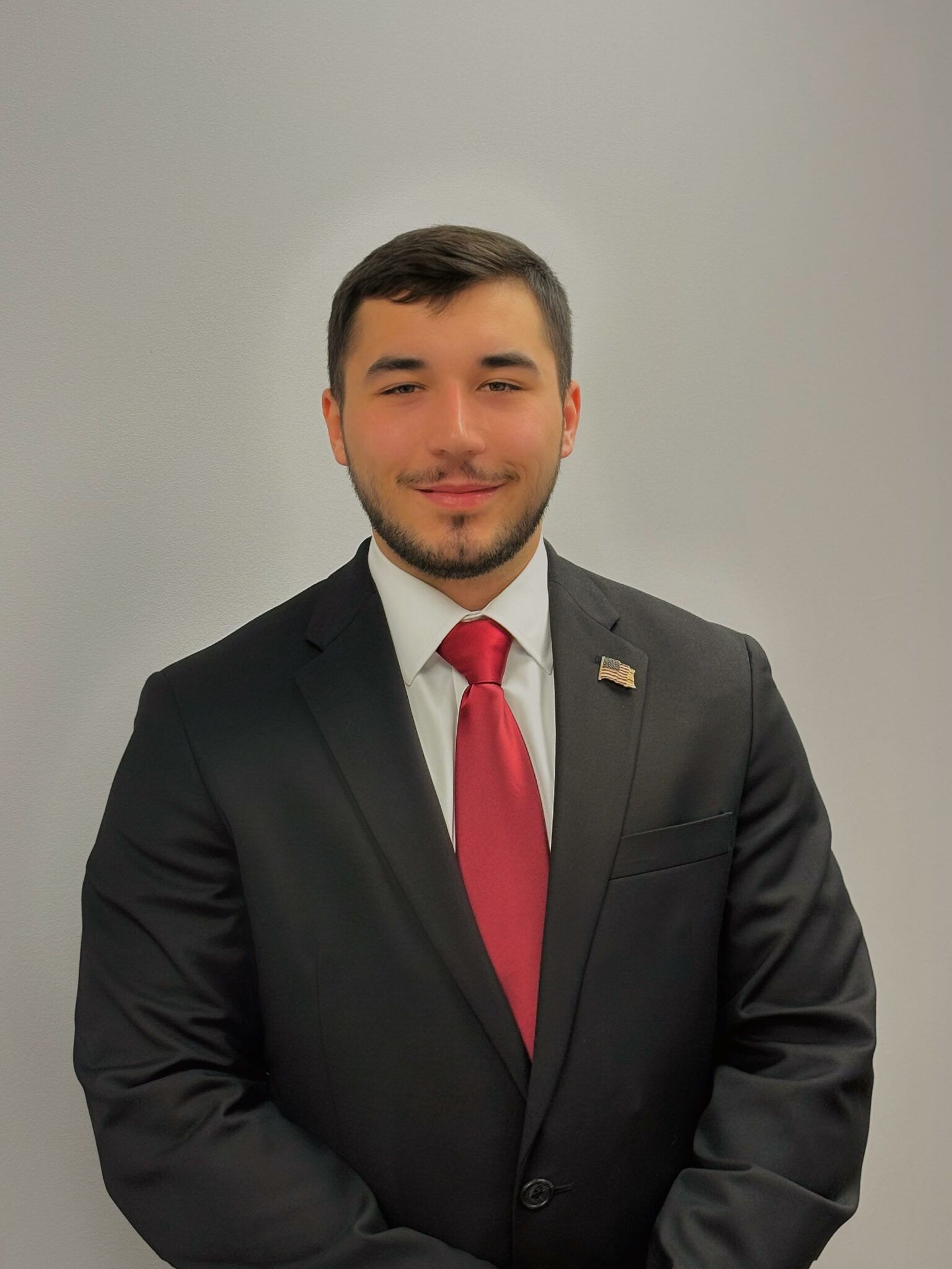 Man in a black suit and red tie standing against a plain gray wall.