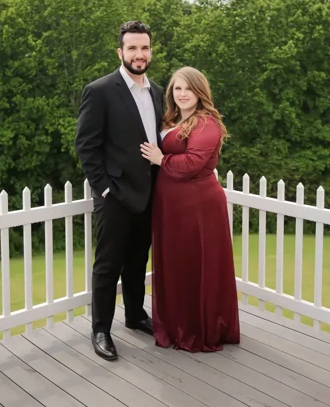 A couple posing on a wooden deck by a white fence, with trees in the background.