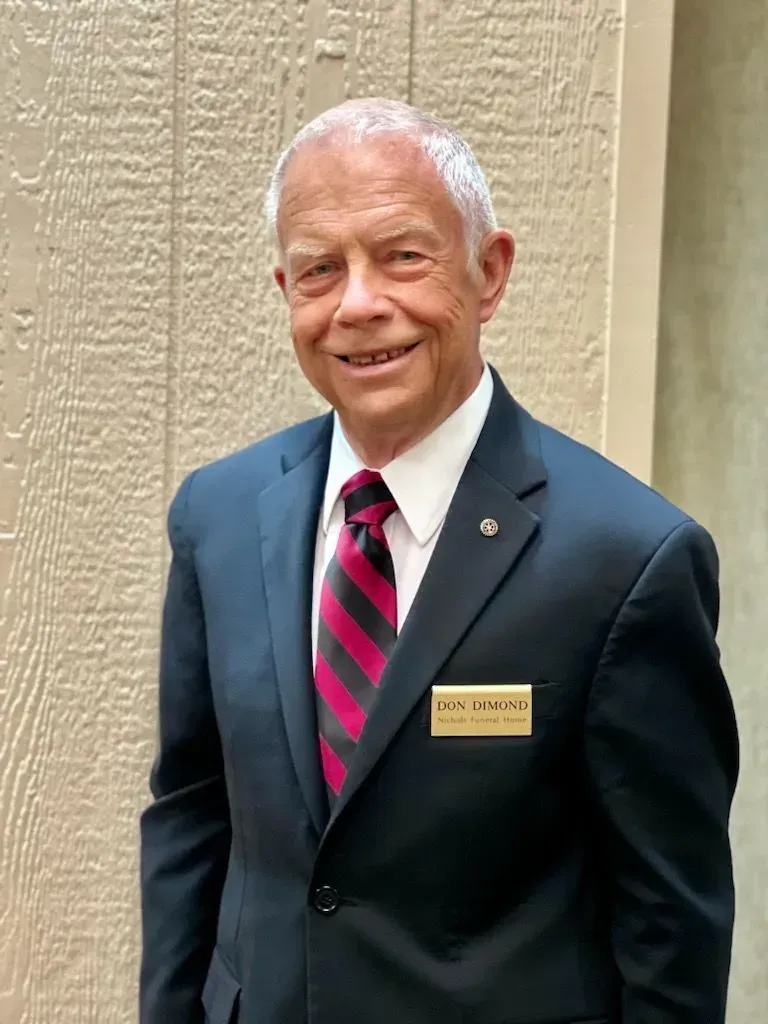Smiling man in a dark suit with pink tie and name badge standing indoors