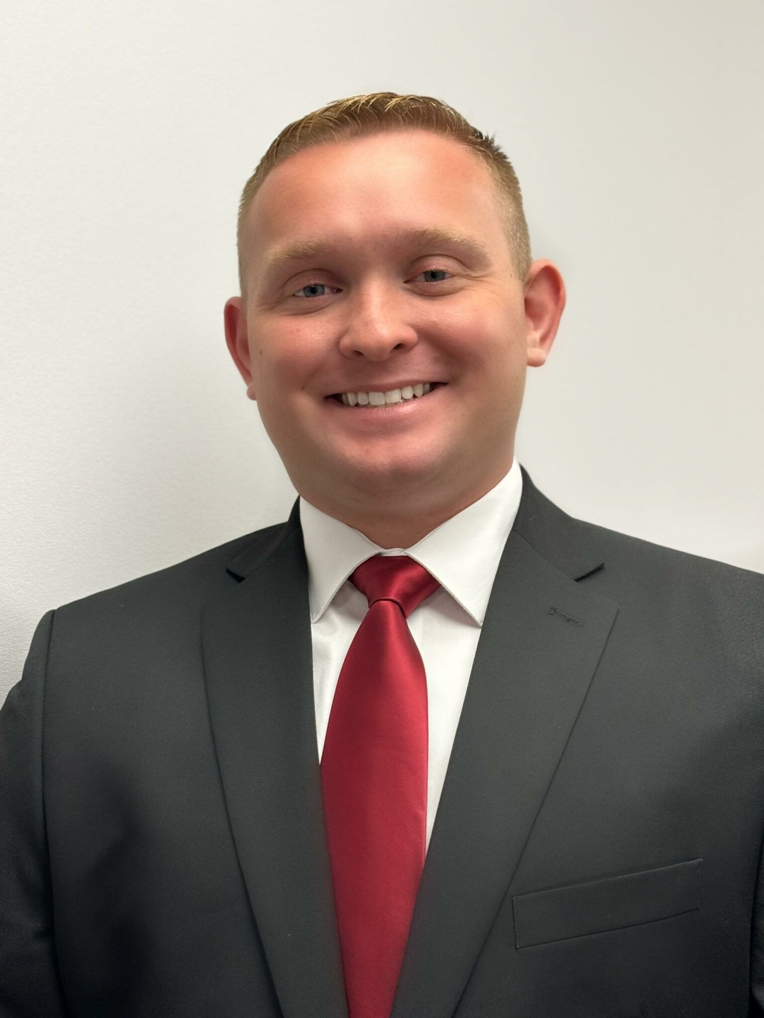 Smiling person in a dark suit, white shirt, and red tie against a plain light background