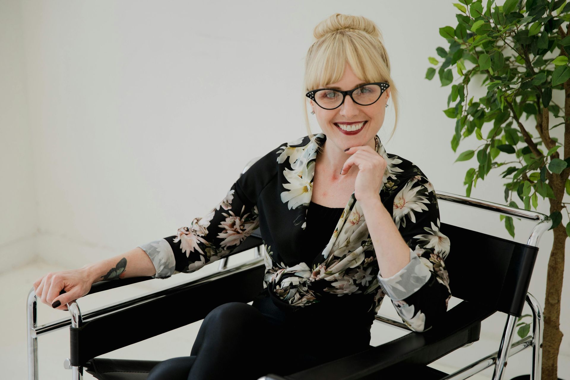 Woman in floral top and glasses, smiling, seated on black chair, near a plant.