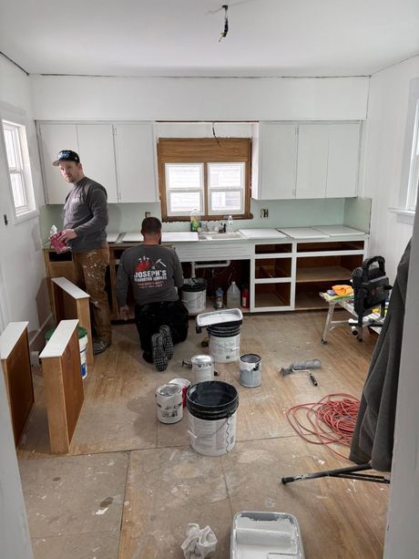 Two workers in a kitchen under renovation, with tools, paint buckets, and unfinished cabinets.