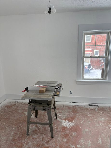 Partially renovated room with a table saw, white walls, bare floor, and a window overlooking brick buildings