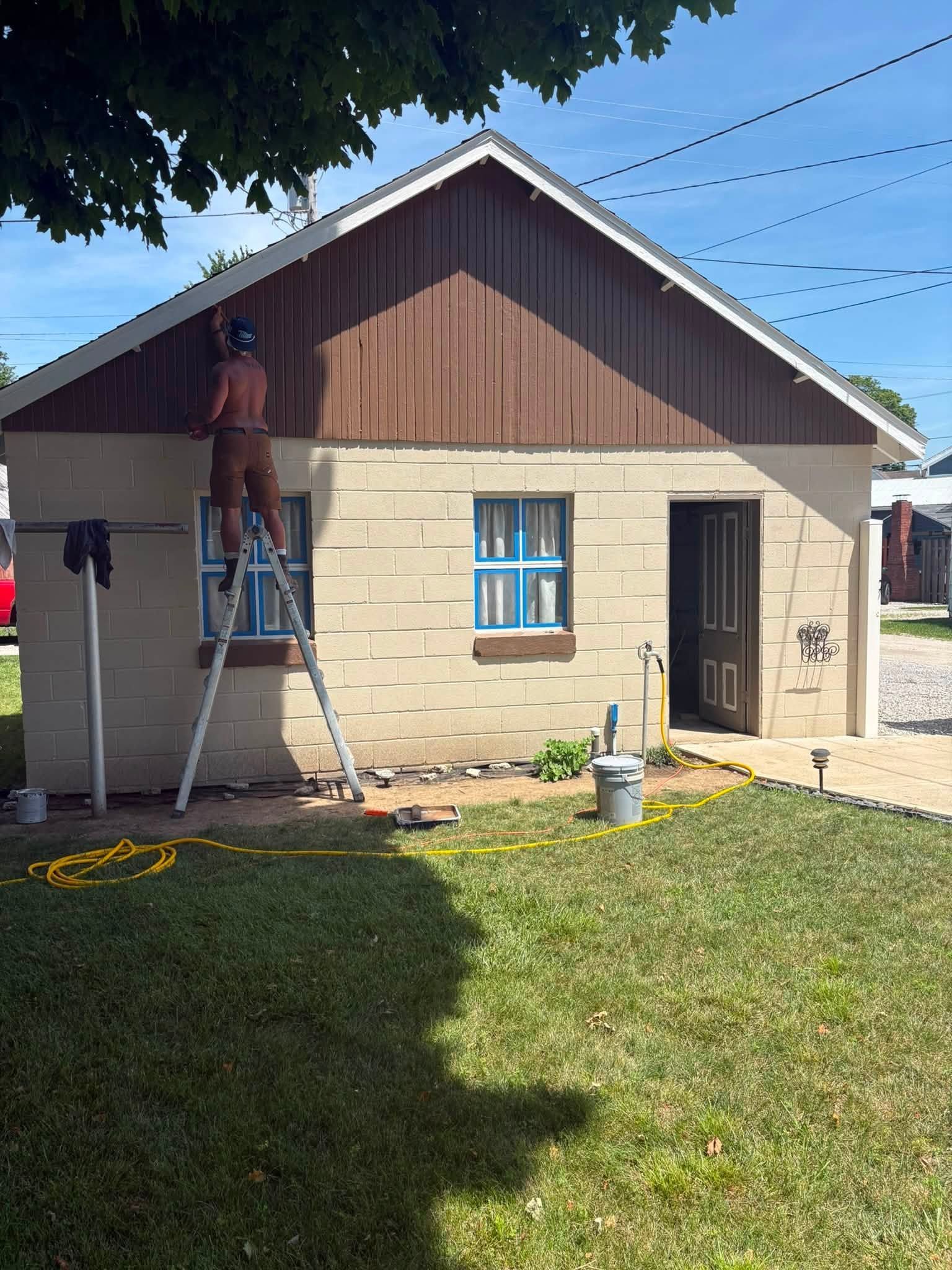 Worker on ladder painting the exterior of a small house with brown siding and beige stone walls.