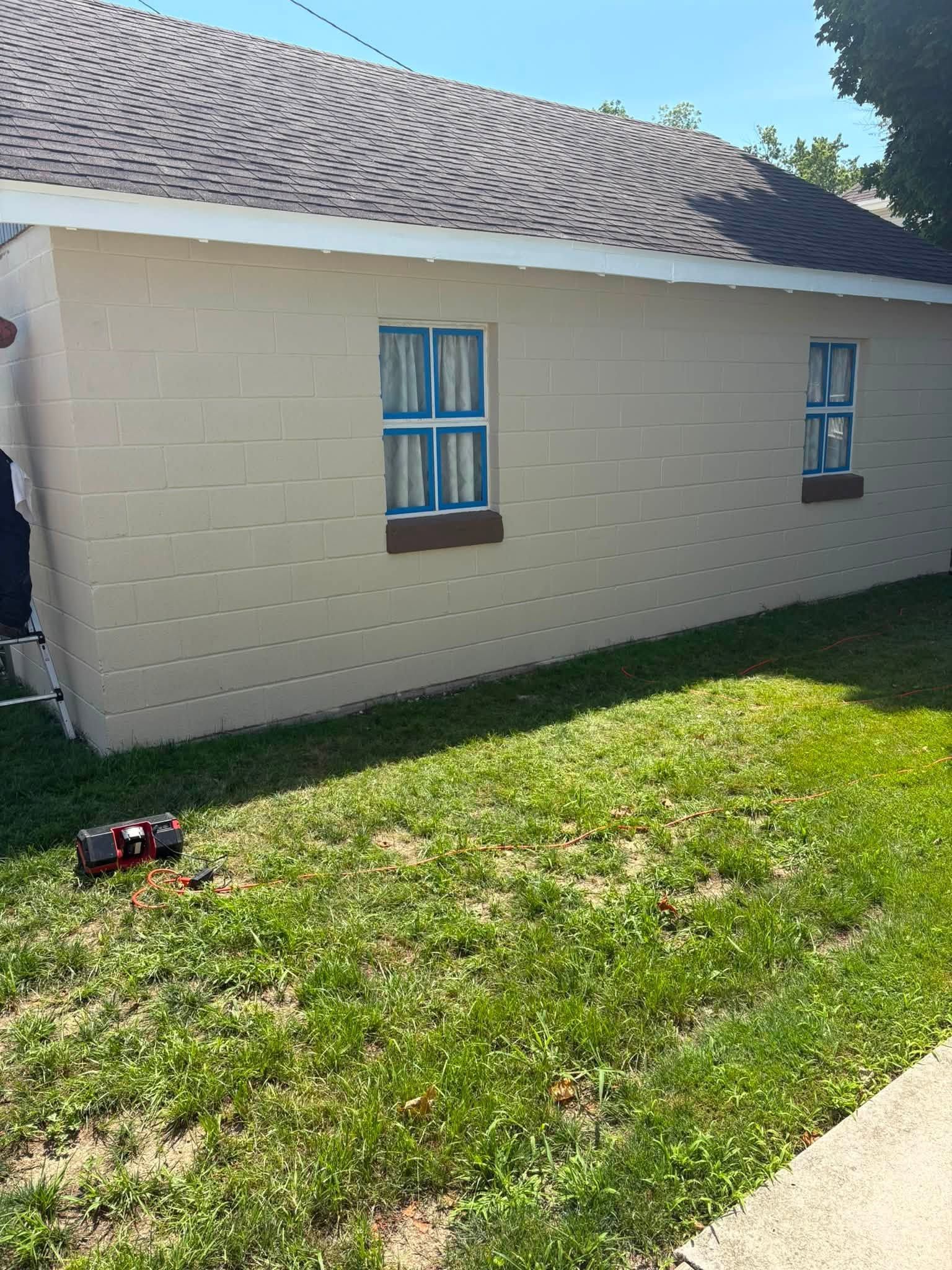 Small beige house with blue-trimmed windows, brown shingled roof, and a grassy yard in sunlight