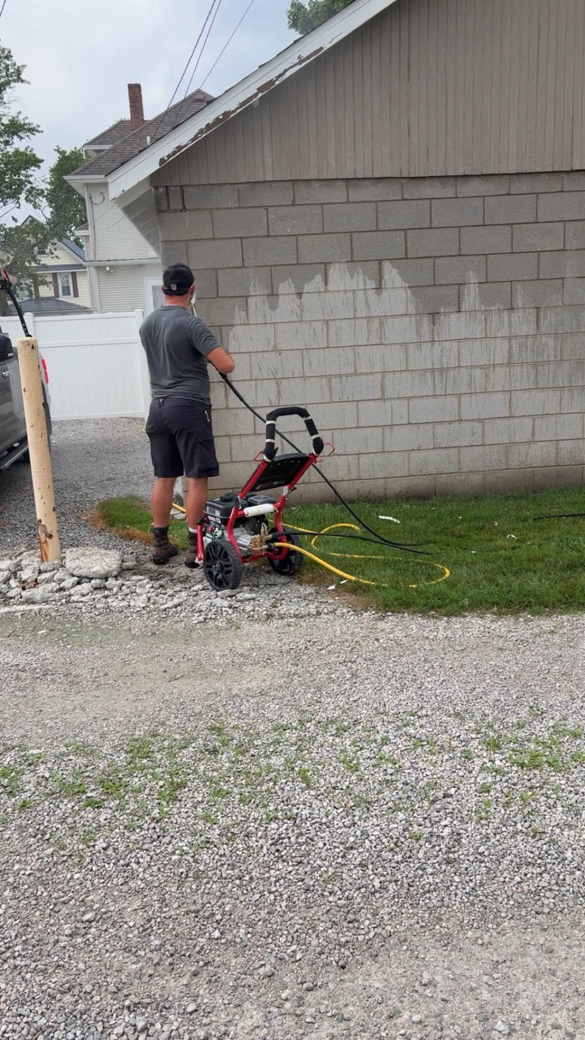 Person power-washing a house wall beside a gravel driveway and lawn