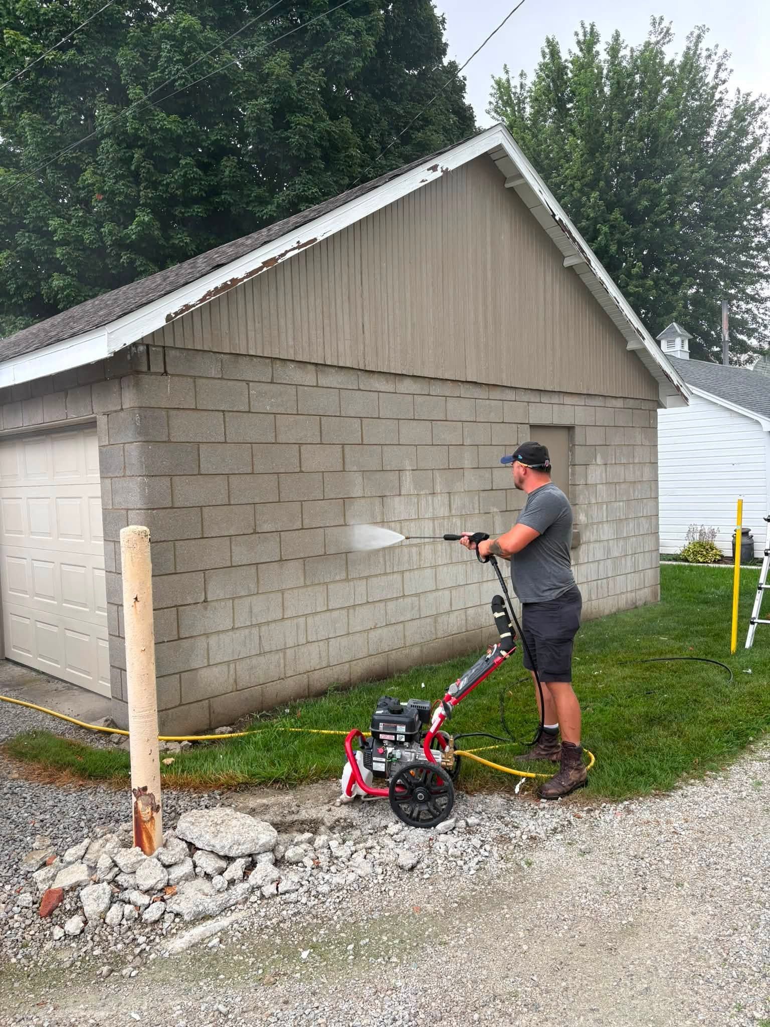 Worker pressure-washing a gray cinderblock garage beside a gravel driveway