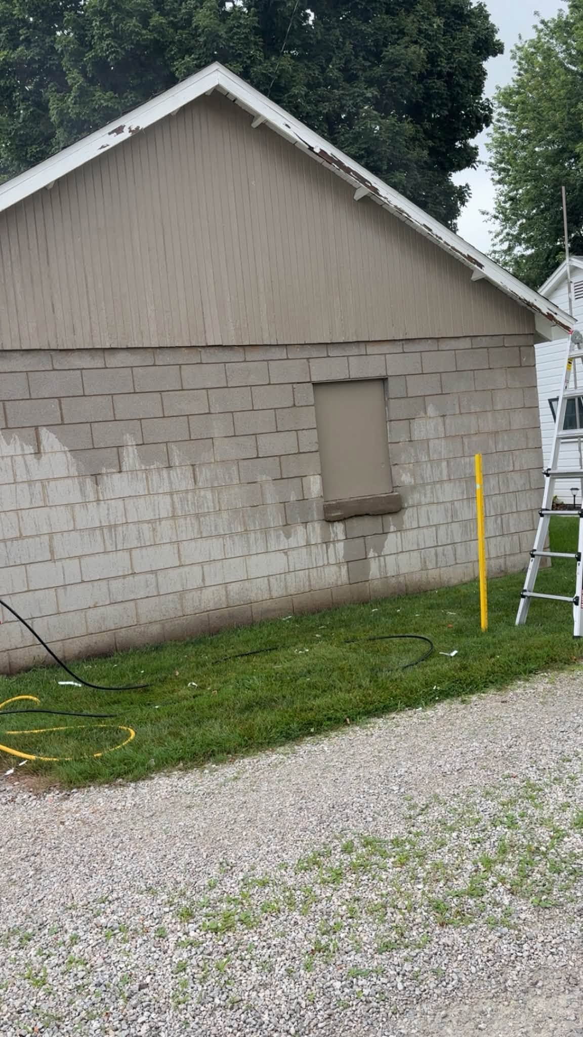 Side of a beige house with a boarded-up window, yellow post, ladder, and gravel driveway.