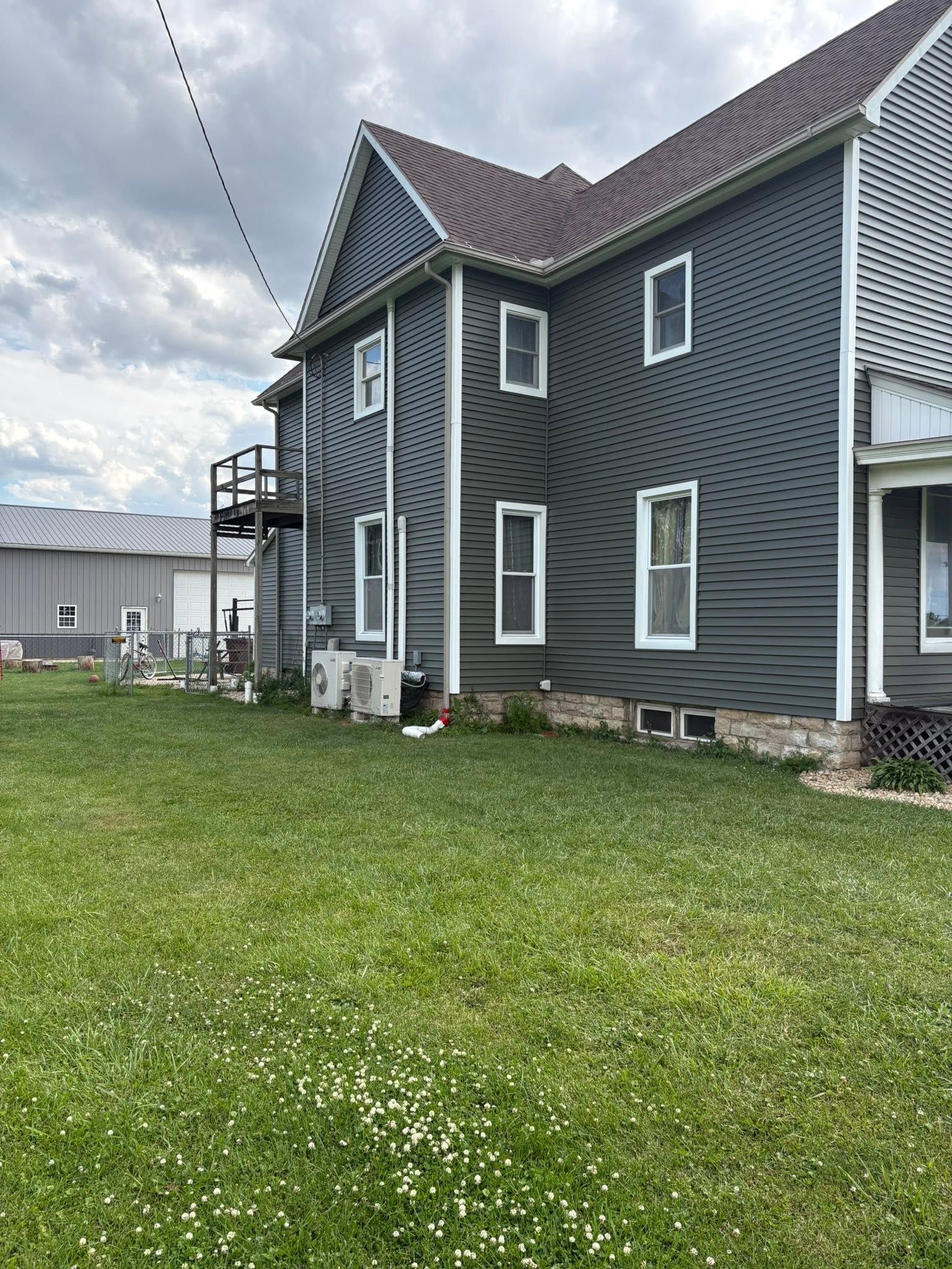 Dark gray house with white trim and lattice siding, side yard with grass and utility units under cloudy sky