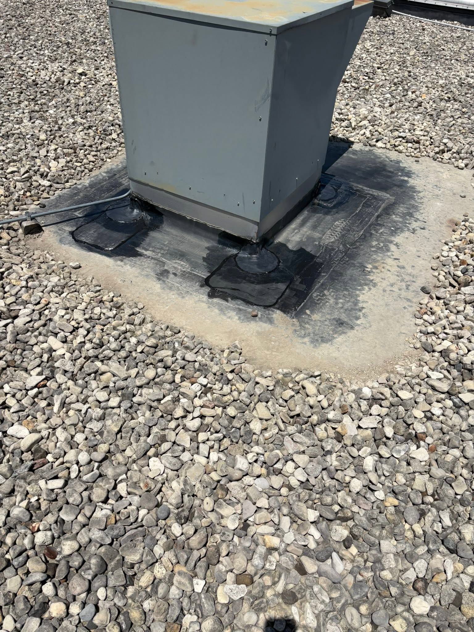 Gray utility box on a gravel roof, mounted on a concrete base with black sealant patches.