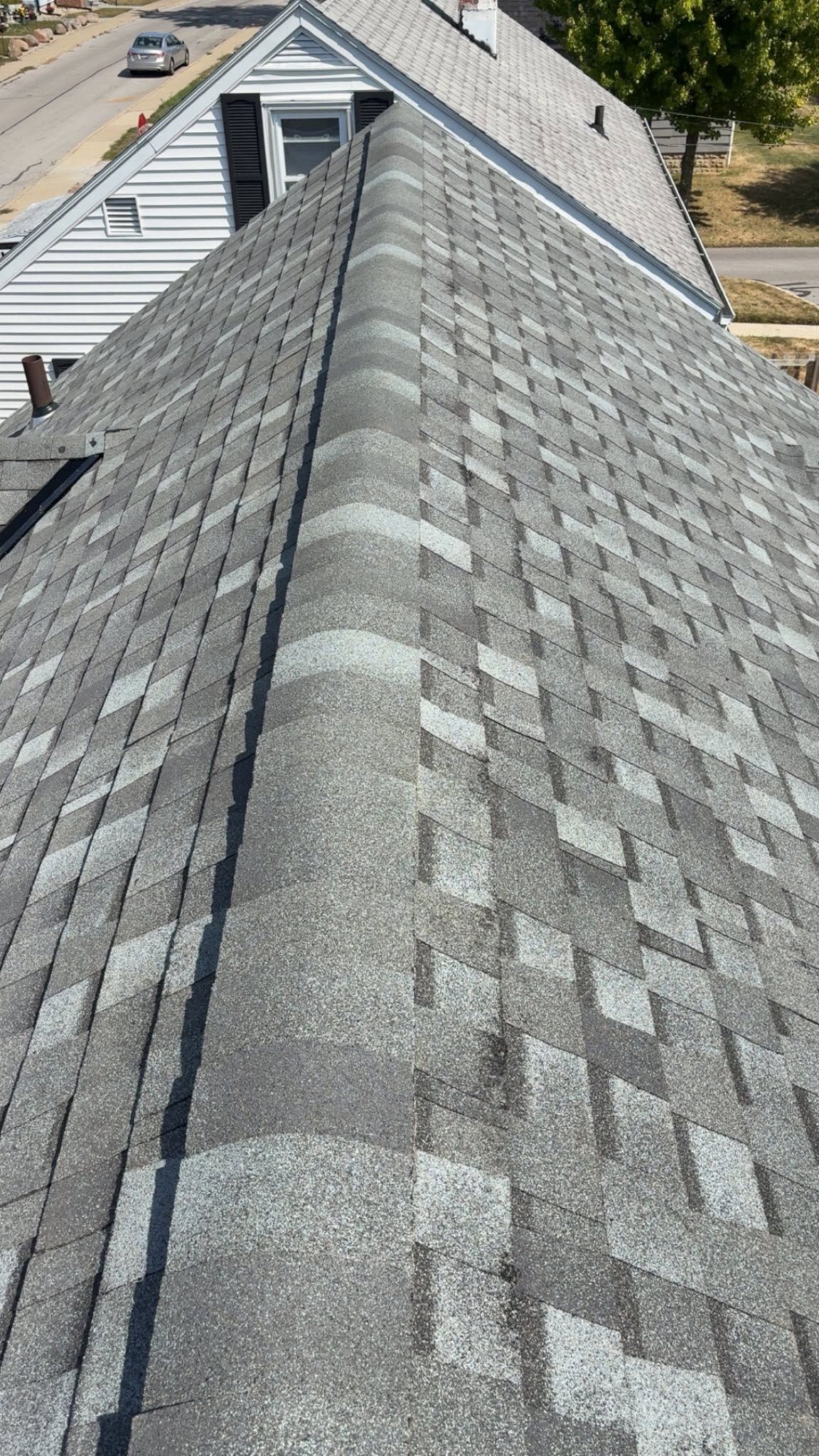 Gray shingled roof with a central ridge viewed from above, with nearby house and trees in the background