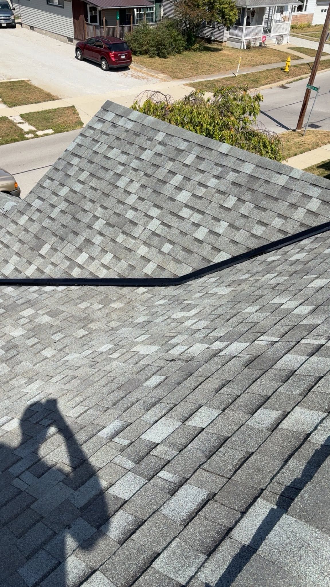 Gray shingled roof with a black ridge line, viewed from above with a street and houses in the background