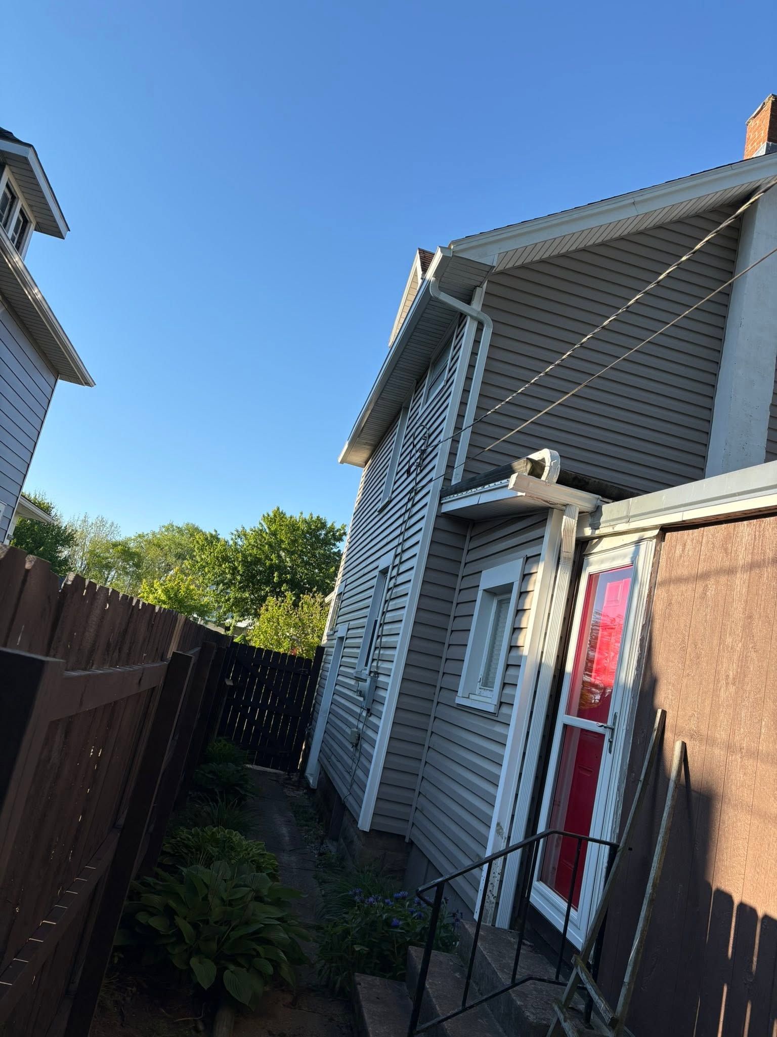 Narrow side yard between houses with a wooden fence, white siding, and a red door under blue sky