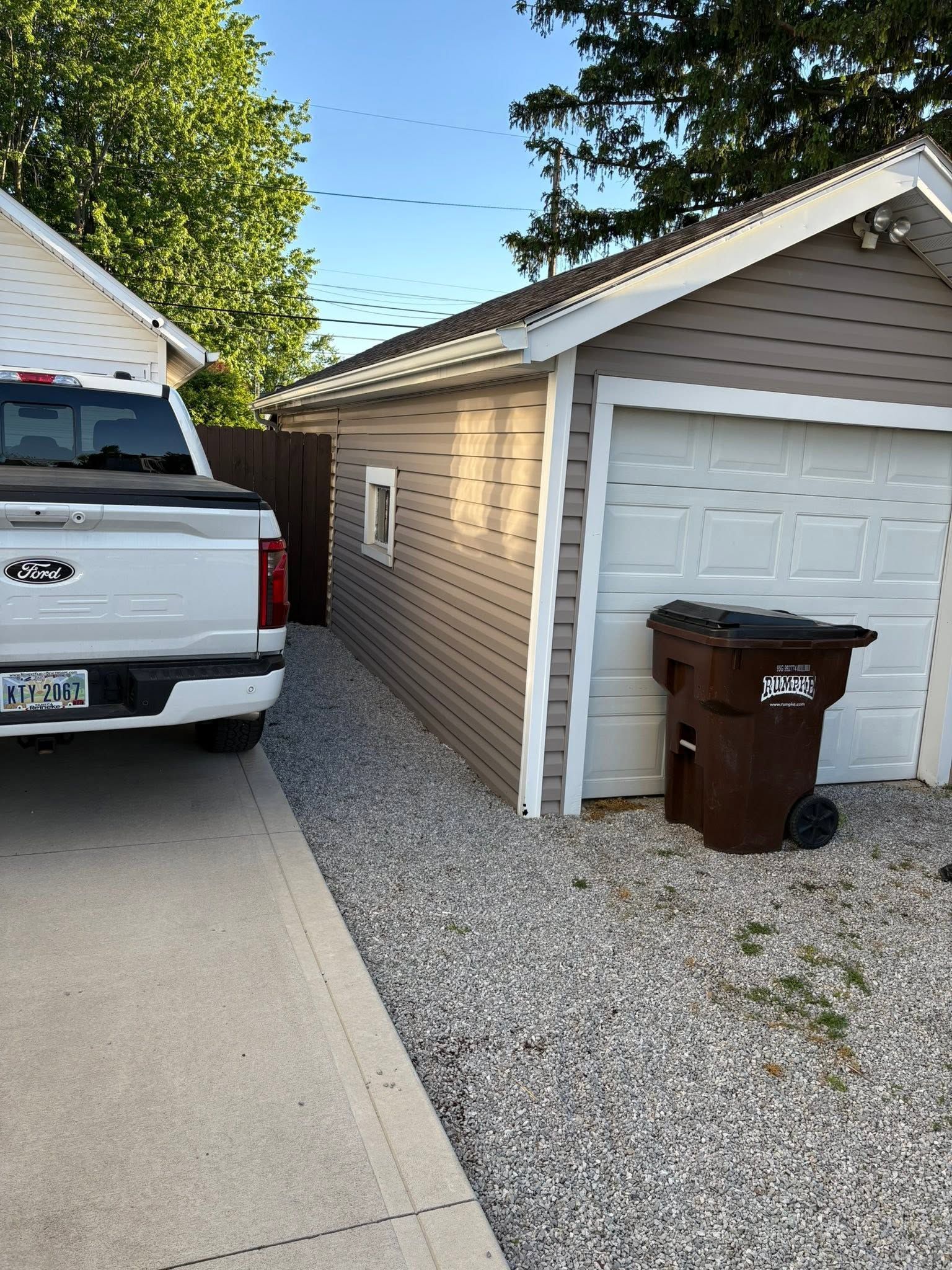 Driveway beside a garage with a white pickup truck and brown trash bin on a gravel strip