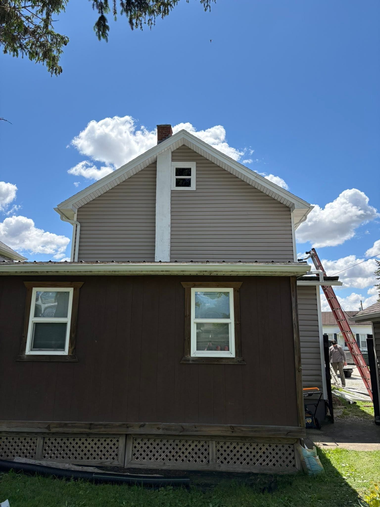 Side view of a house with brown siding, beige upper gable, and a blue sky with clouds overhead.