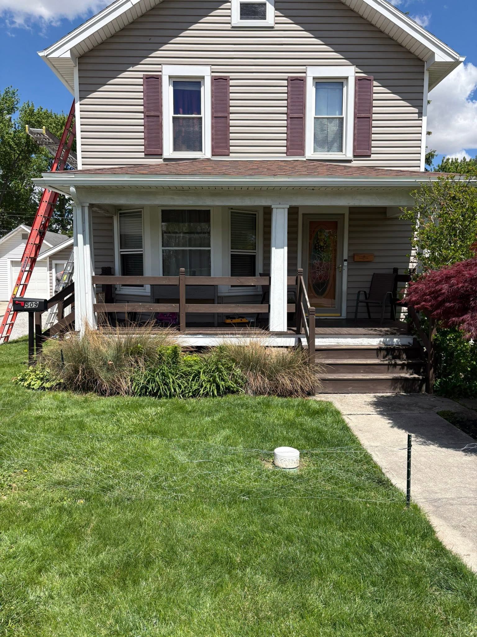 Two-story beige house with front porch, white trim, red shutters, and a green lawn in front