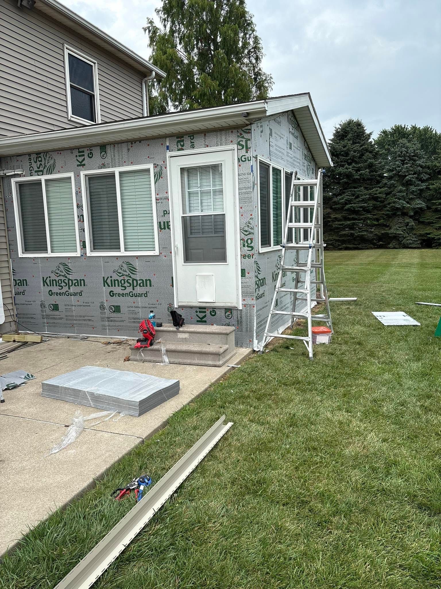 Small house porch with white siding, windows, steps, and construction materials in a grassy yard.