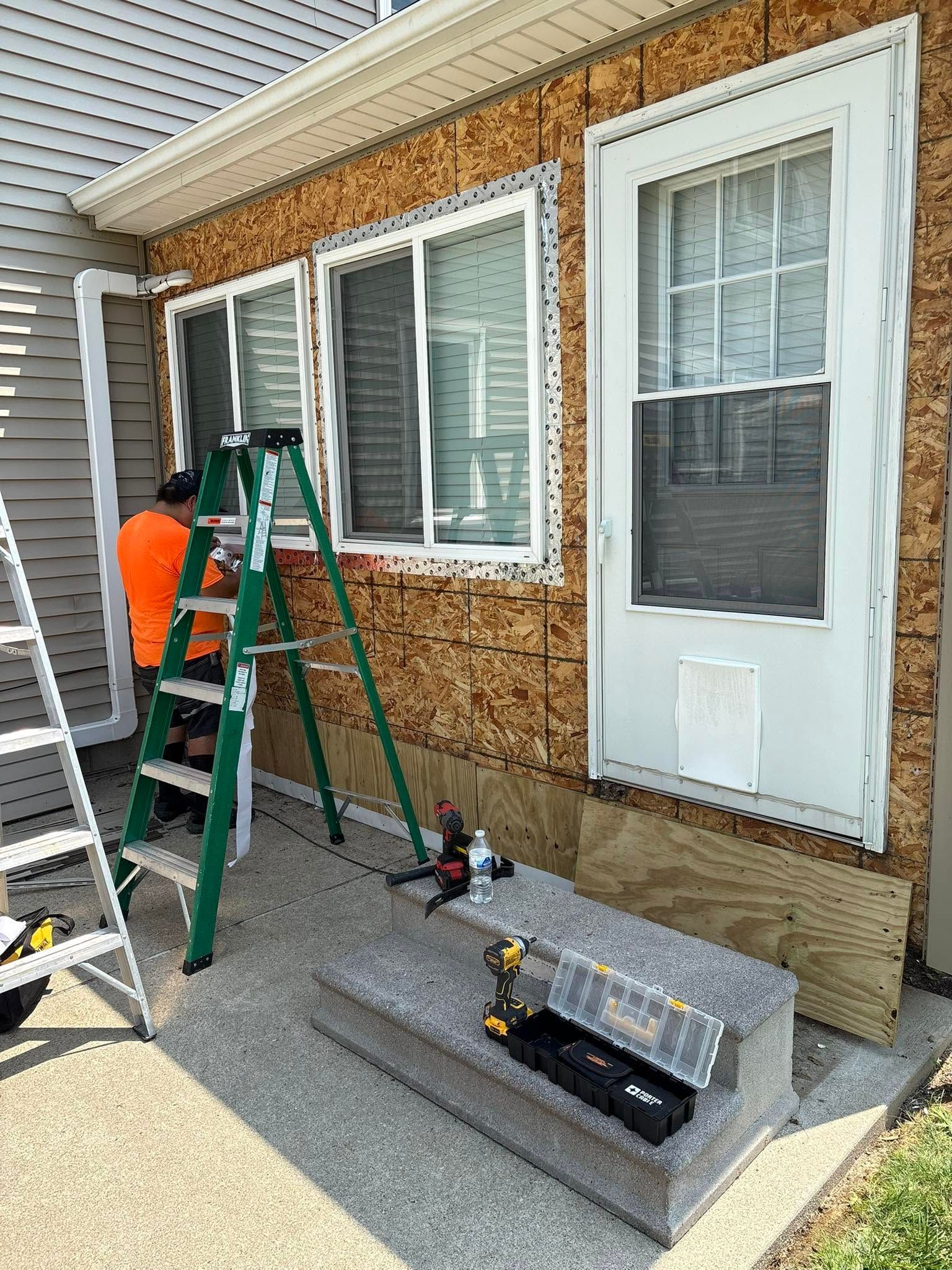 Worker on ladder installing siding on a brick house exterior, with tools on the sidewalk.