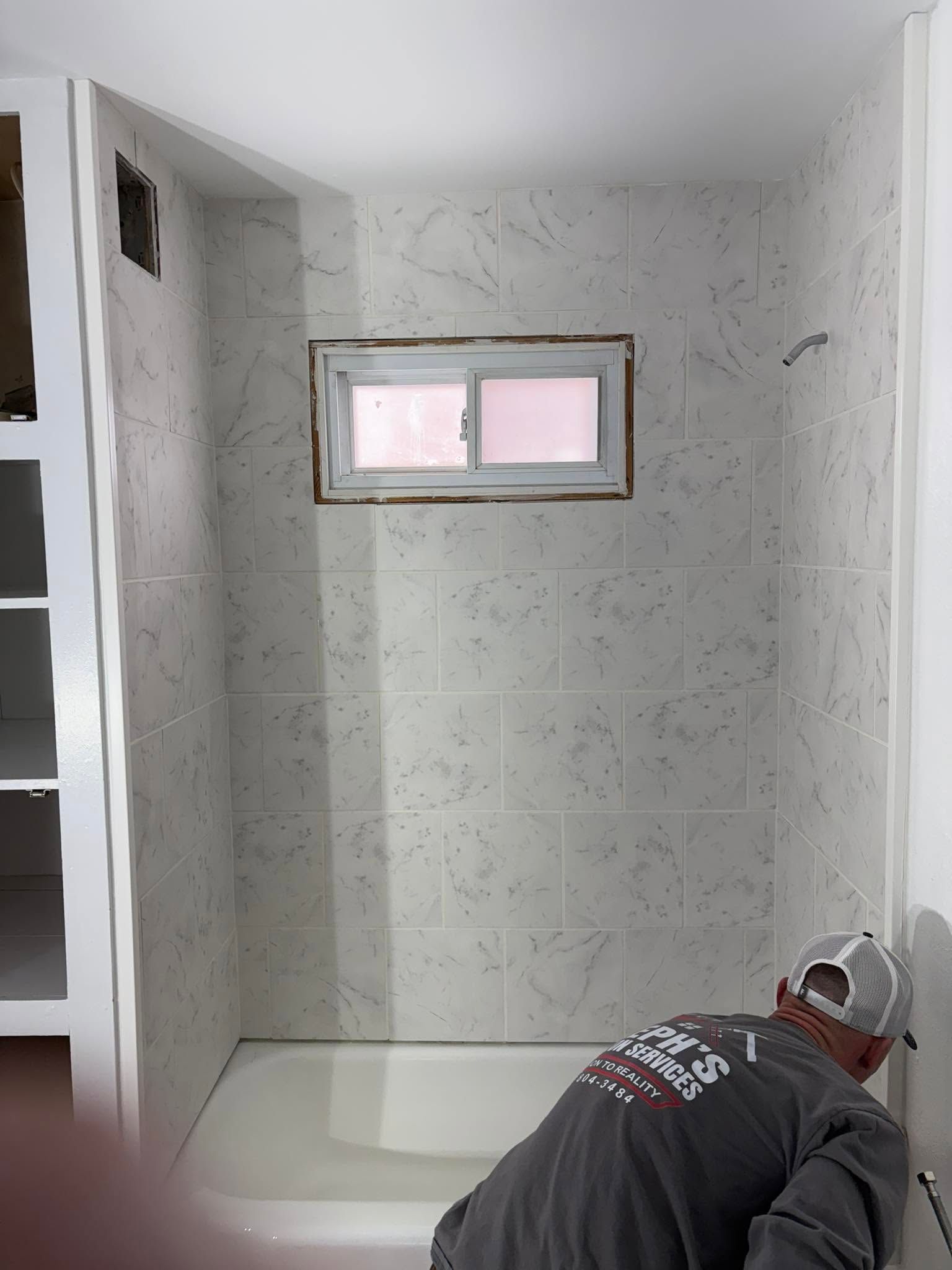 Worker tiling a small white bathroom shower with a pink-tinted window above the tub.