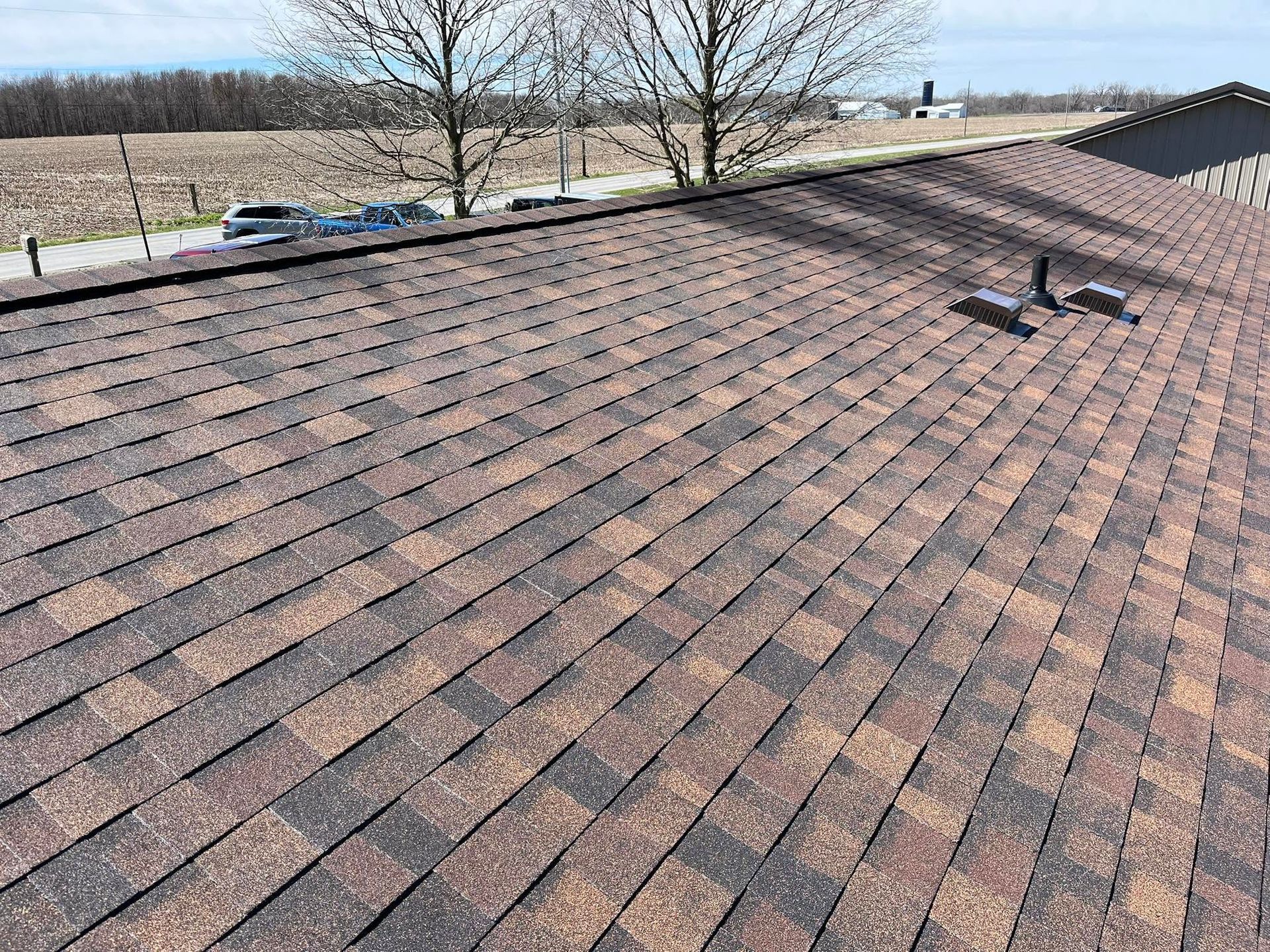 Shingled rooftop with vent pipes overlooking fields and leafless trees under a clear sky