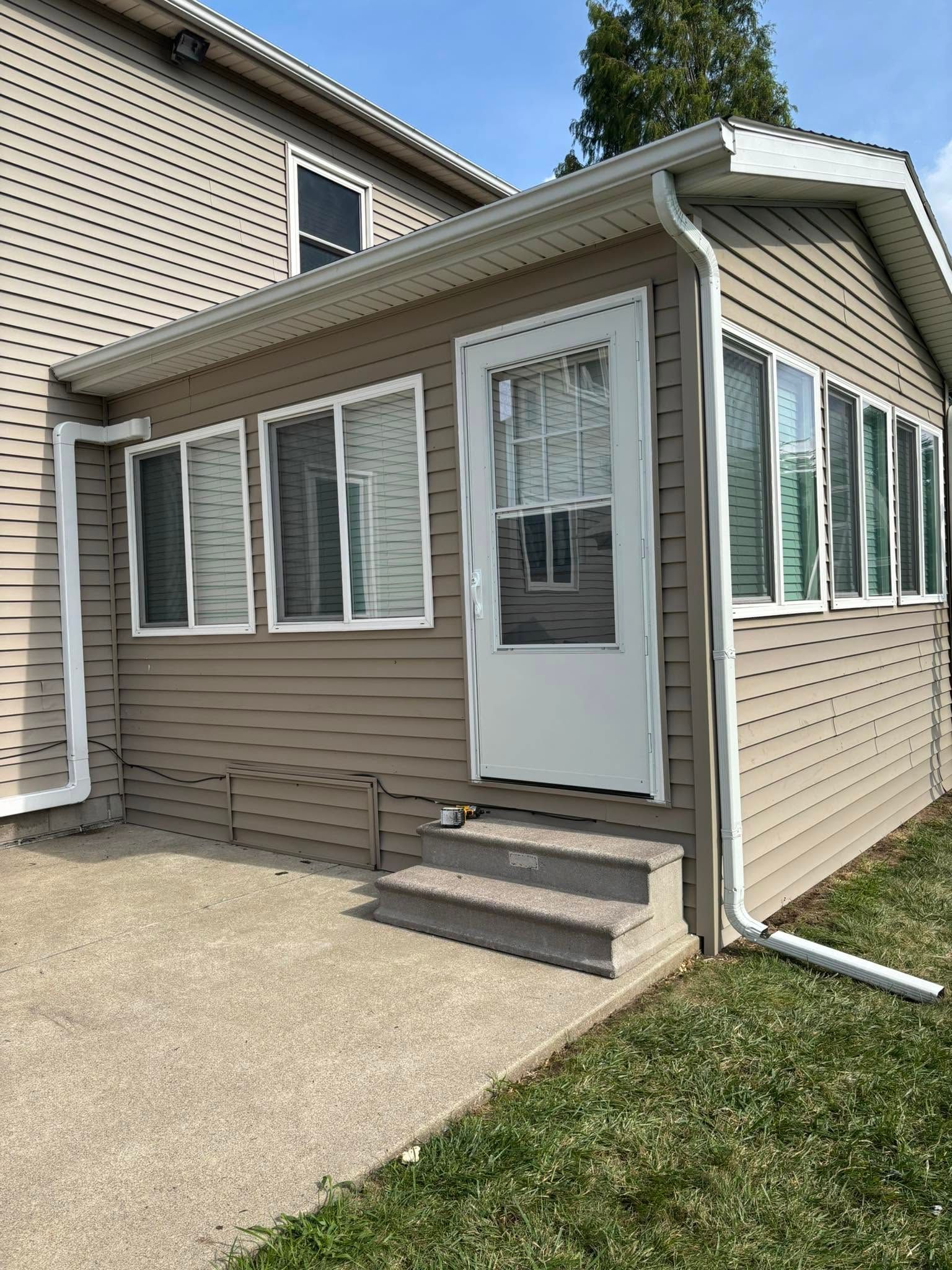Small beige house entrance with white door, windows, steps, and concrete walkway beside a lawn.