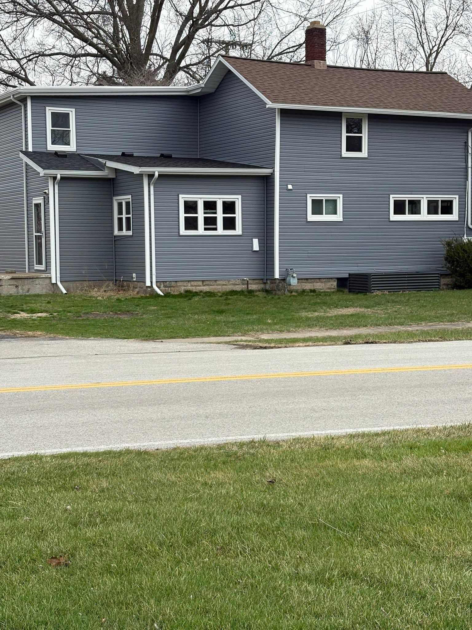 Gray house with white trim and a porch beside a road, seen from across a grassy lawn.