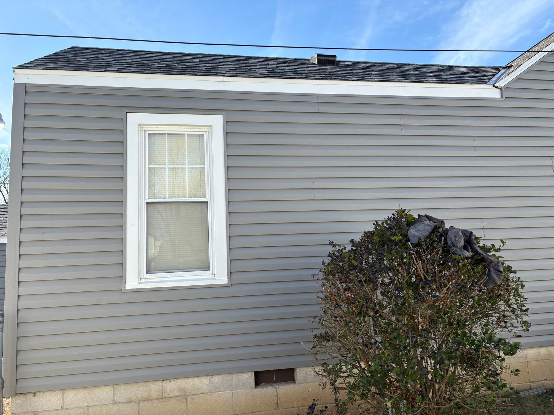 Gray house siding with a small white-trimmed window and a bush in front under a blue sky