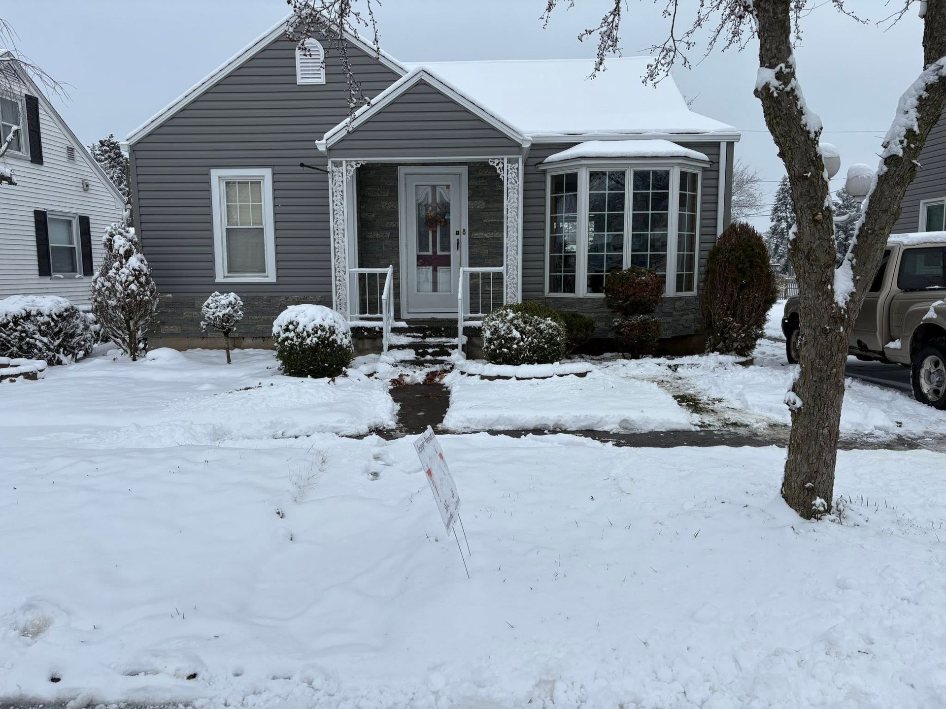 Snow-covered gray house with a front porch and bare trees in the yard