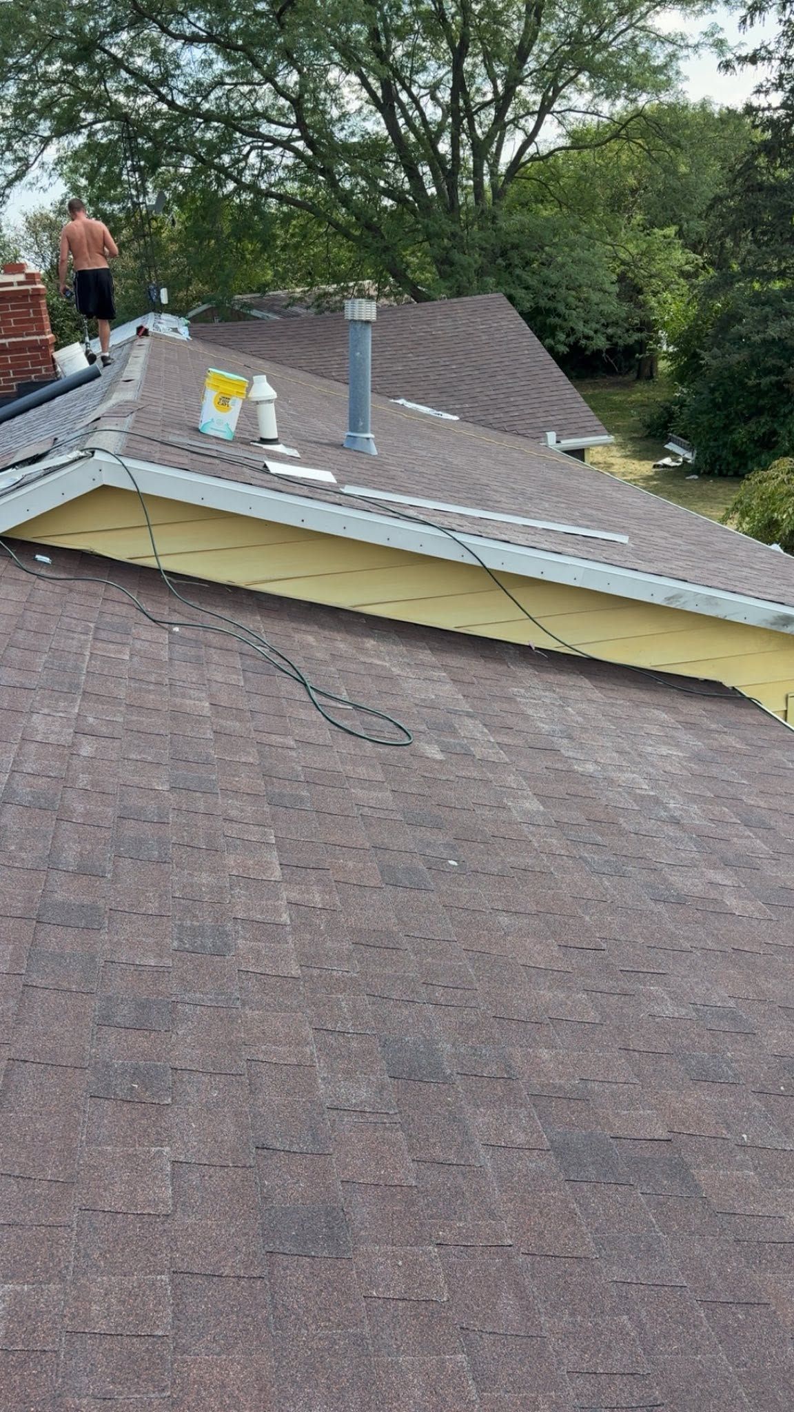 Roof with brown shingles, yellow flashing, and a gray vent pipe, with a person on the roof near trees.