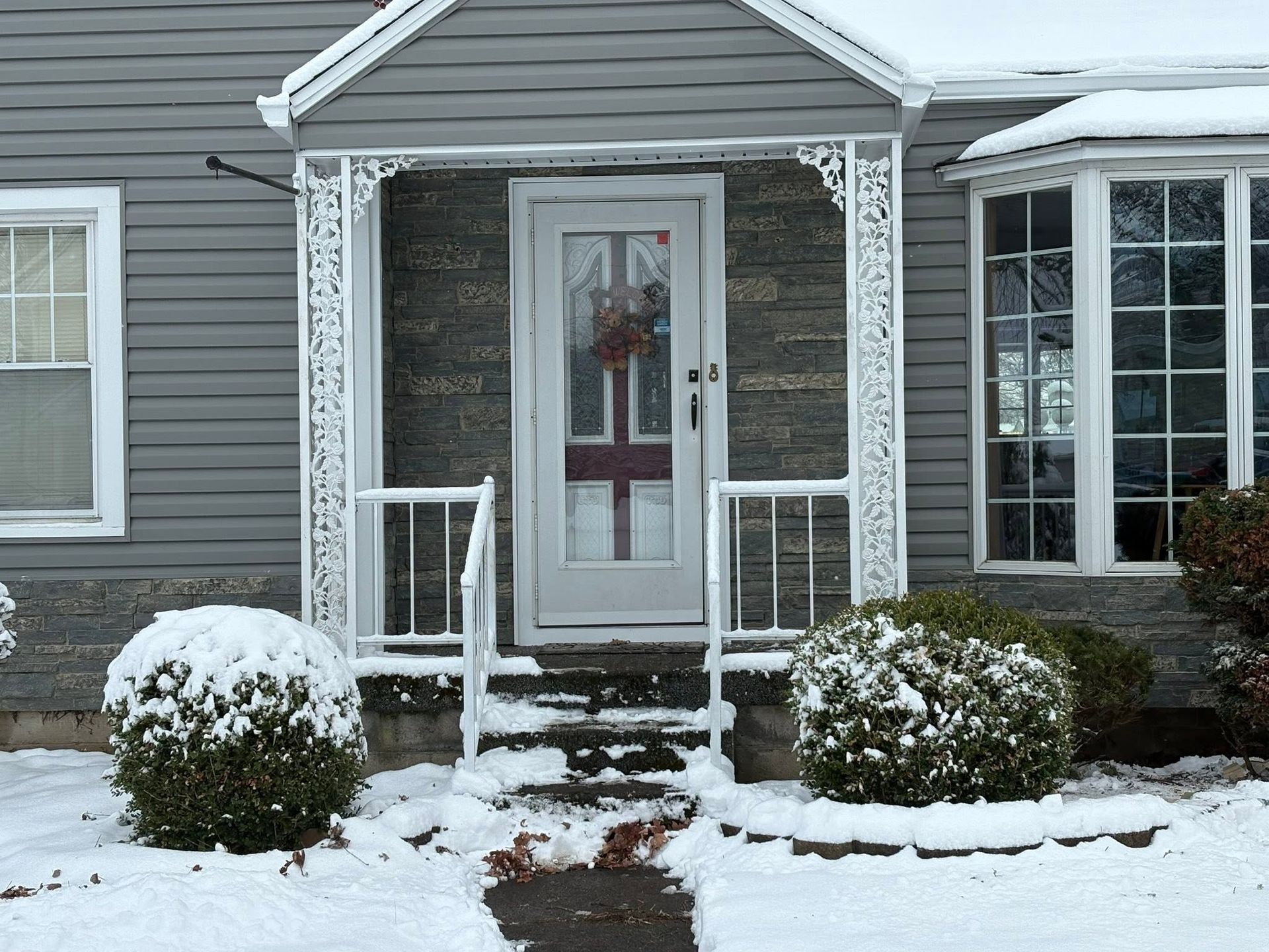 Snow-covered front porch of a gray house with a glass door and white railings.