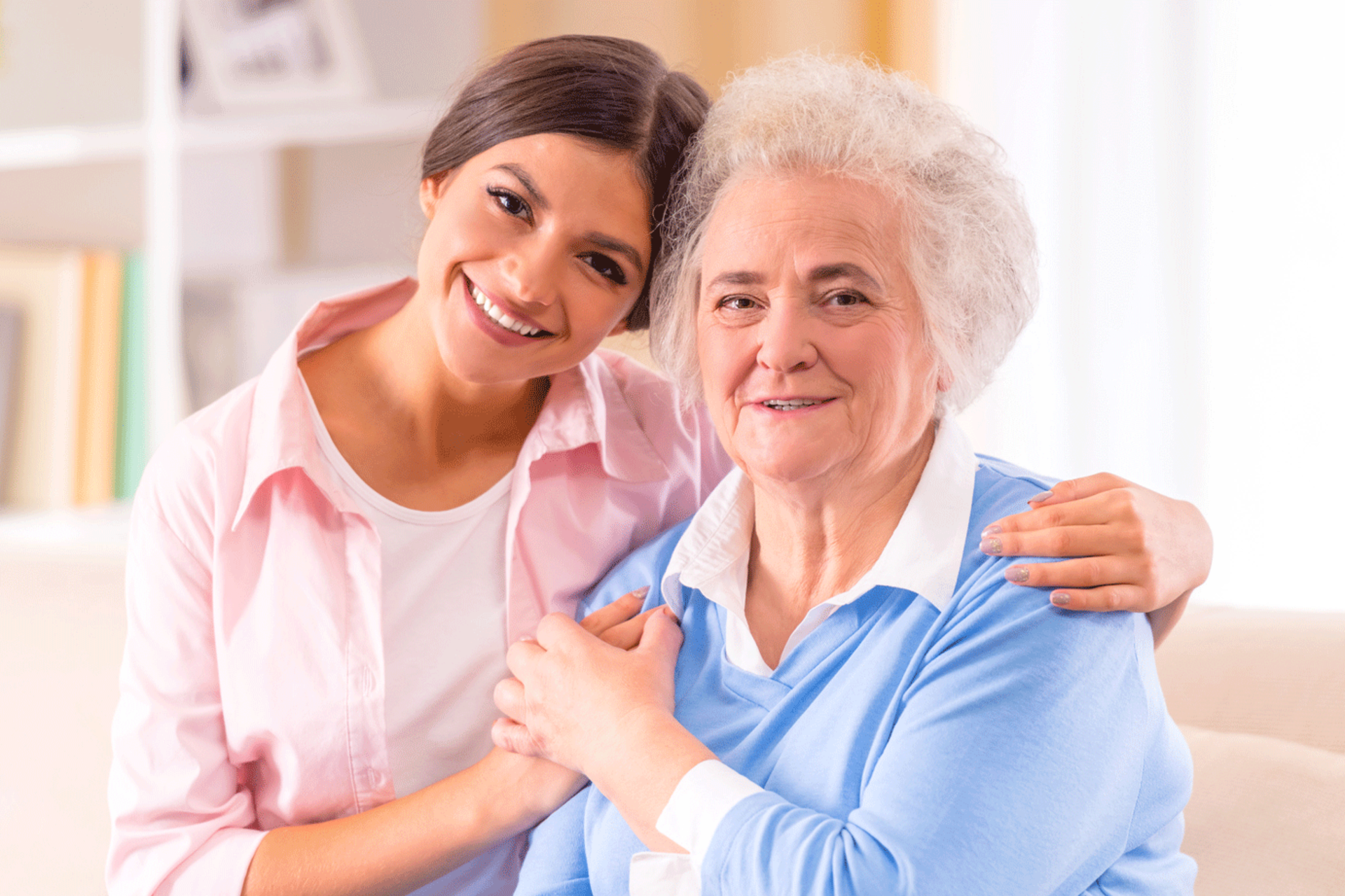 Woman embraces an older woman, both smiling. The younger woman wears pink, older woman blue. Indoors.
