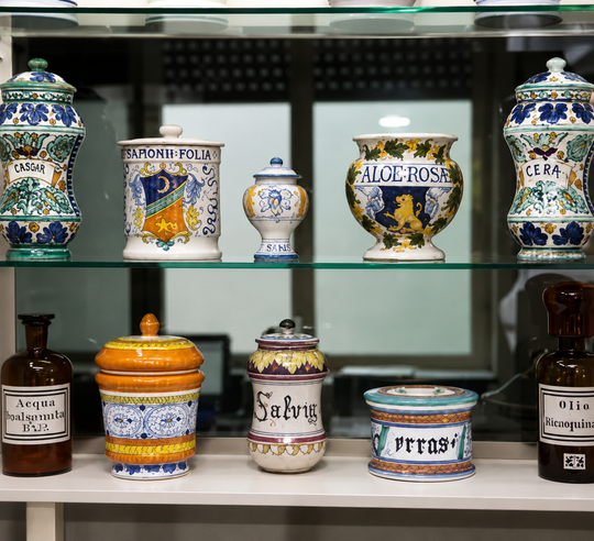 Una vetrina che espone su due ripiani un assortimento di vasi decorativi in ceramica e contenitori da farmacia.