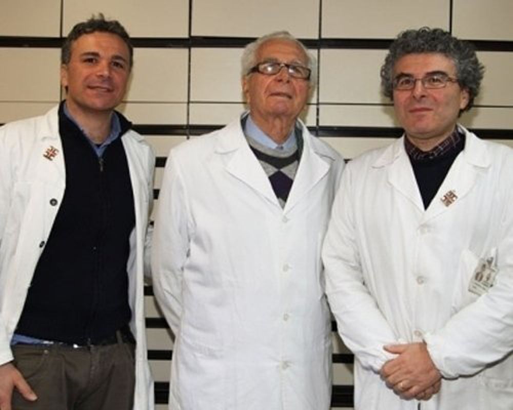 Tre uomini in camice bianco, in piedi uno accanto all'altro in un ambiente interno, rivolti verso la telecamera.