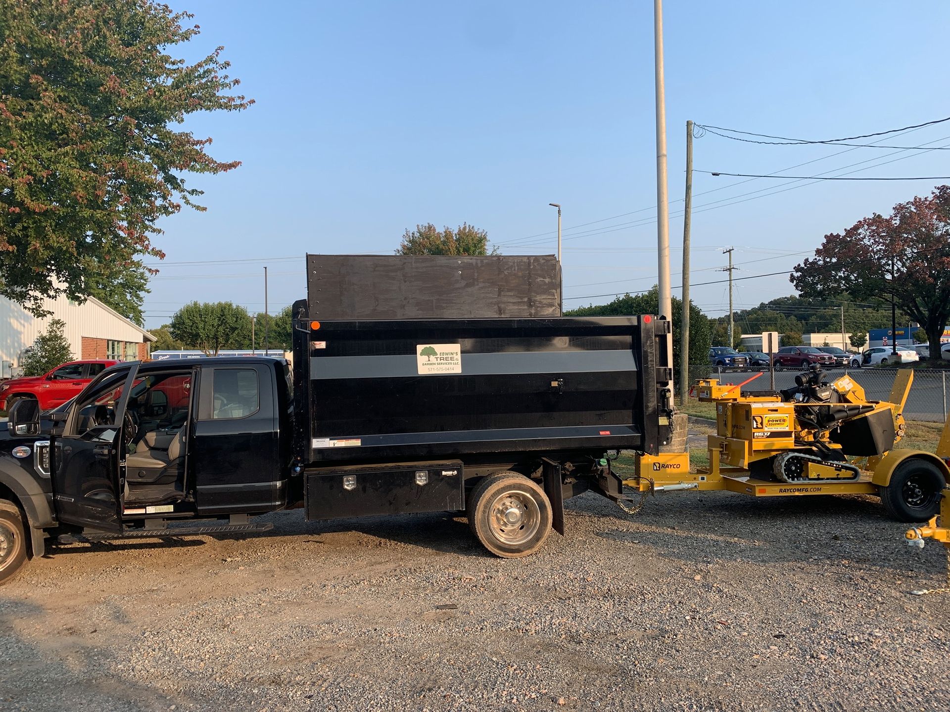 Black dump truck with trailer hitched to a yellow wood chipper on a gravel lot near a building.