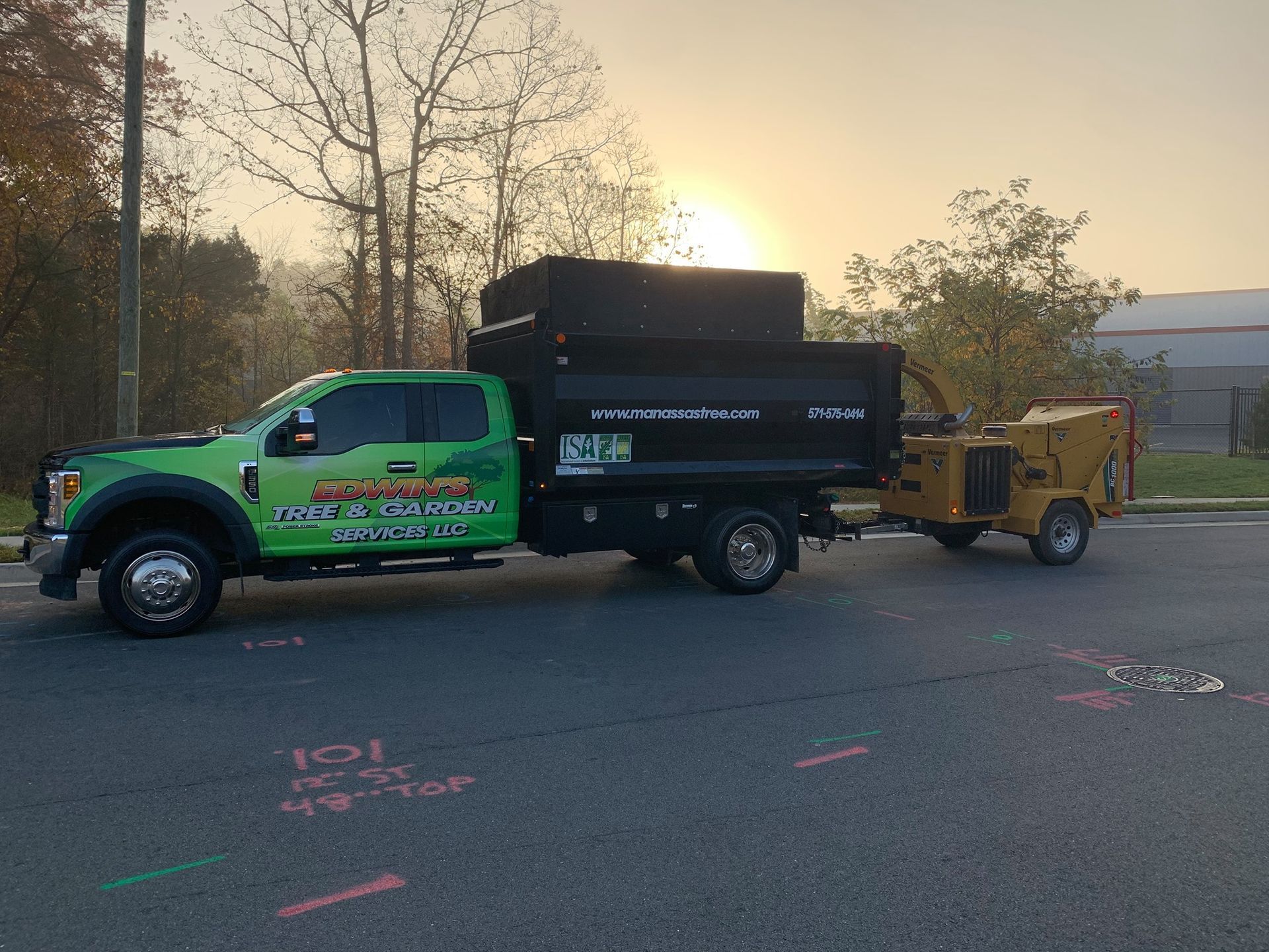 Green landscaping truck with a wood chipper trailer parked in a lot, with the sun setting.