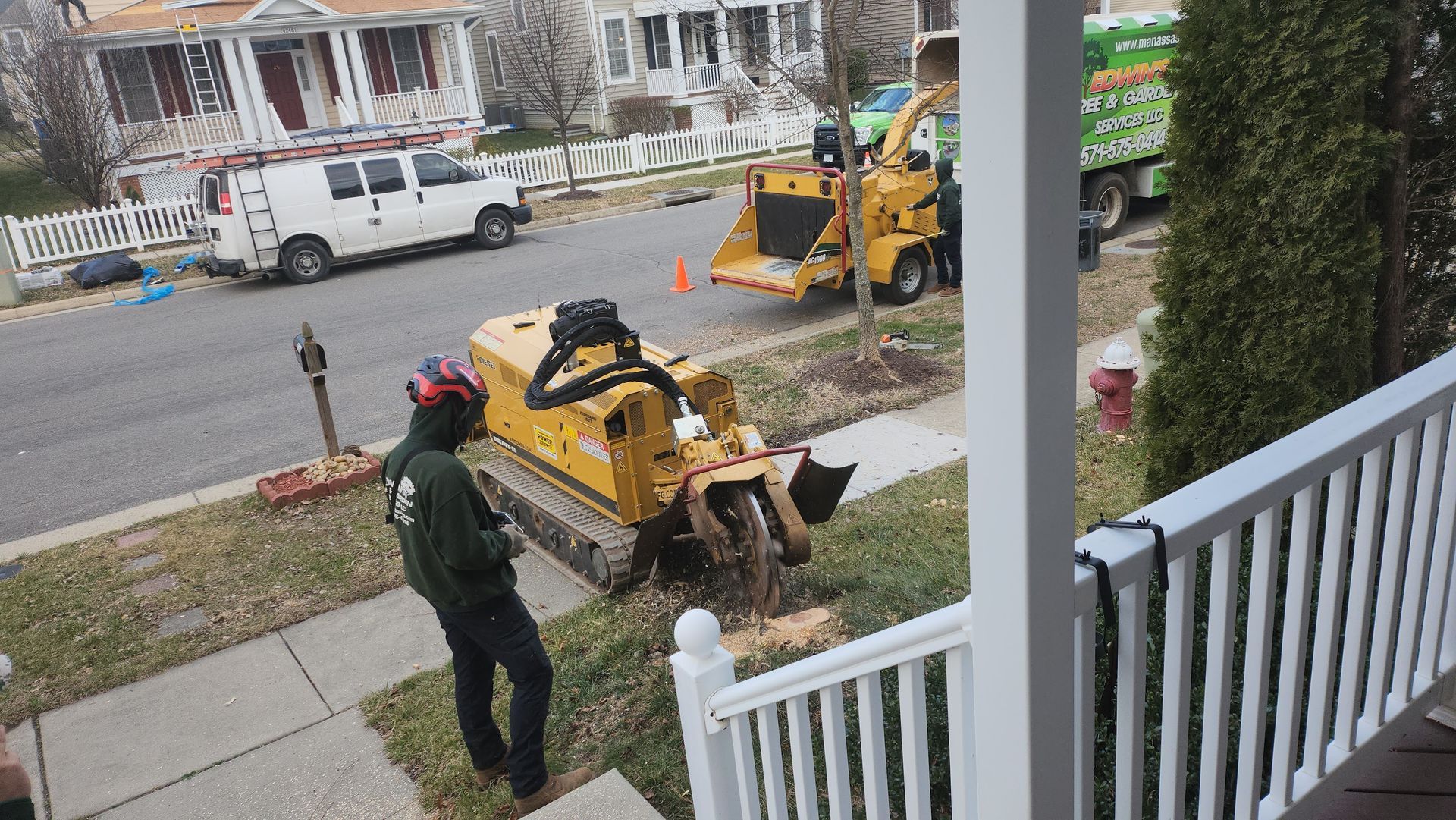 Man operating a stump grinder on a residential street. A truck and other equipment are nearby.